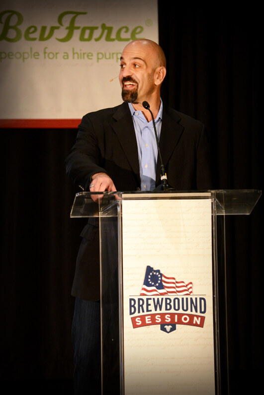 A bald, goateed man in a dark blazer and light blue shirt with a headset microphone smiles at a clear podium marked Brewbound Session, beneath a BevForce banner.