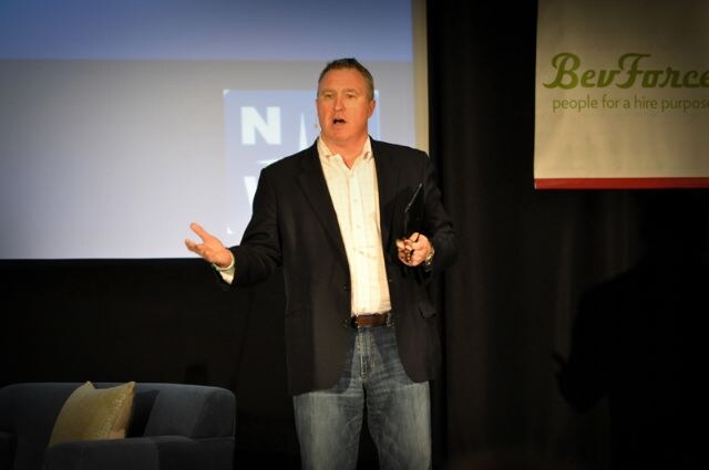 A man in a dark blazer, white shirt and jeans gestures while speaking on stage. A BevForce banner hangs at right and a blue armchair sits at left.