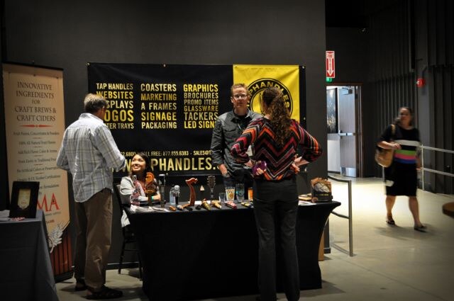 Two men and a woman talk at the Taphandles booth, which has a black banner listing tap handles, coasters and signage. A seated woman is behind the table and a passerby walks at right.