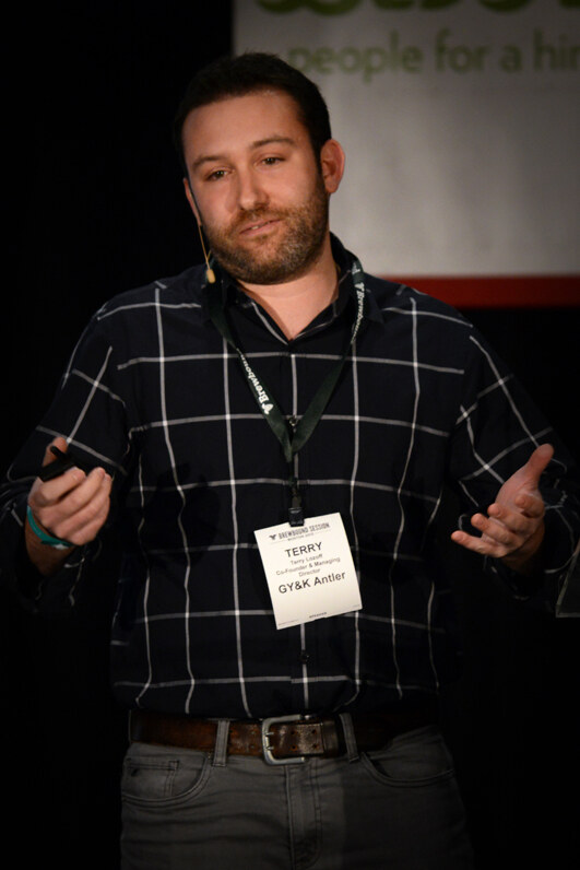 Terry of GY&K Antler, a bearded man in a dark plaid shirt with a headset microphone and badge, gestures with both hands while speaking on stage.
