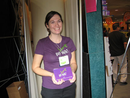 A smiling woman with dark hair wears a purple Hemp got milk? t-shirt and a name badge. She holds a purple Hemp got milk? carton in front of a curtained booth entrance.