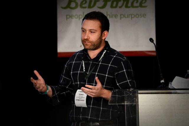 Terry of GY&K Antler, a bearded man in a dark plaid shirt with a badge and lanyard, gestures with both hands while speaking at a podium in front of a BevForce banner.