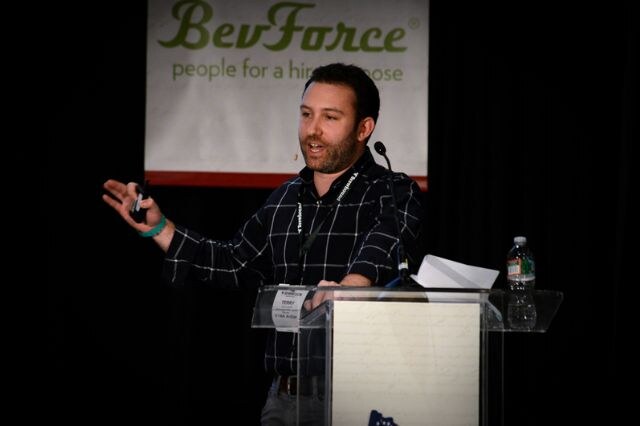Terry, a bearded man in a dark plaid shirt, speaks at a clear podium and gestures with a clicker in one hand. A BevForce banner hangs behind him, and a water bottle sits on the podium.