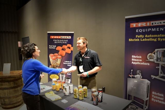 Two people shake hands at the TRIpack booth, a man in a black polo with a lanyard and a woman in a blue sweater. Canned samples sit on the table before TRIpack banners.