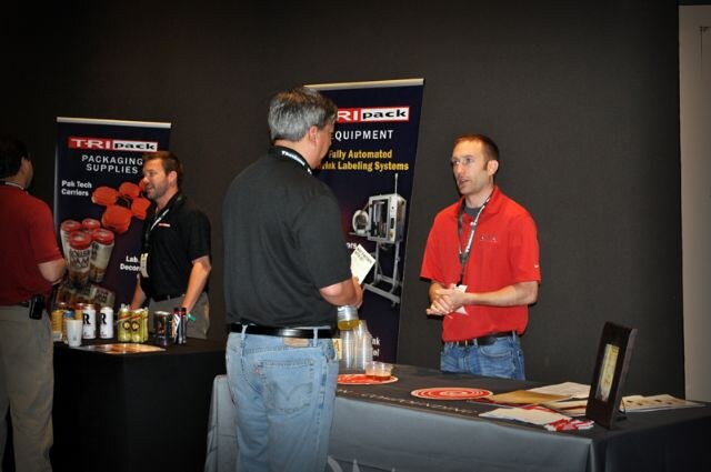 A man in a red polo talks with a man in a black shirt facing away at the TRIpack booth. Another man stands behind the table near TRIpack banners.
