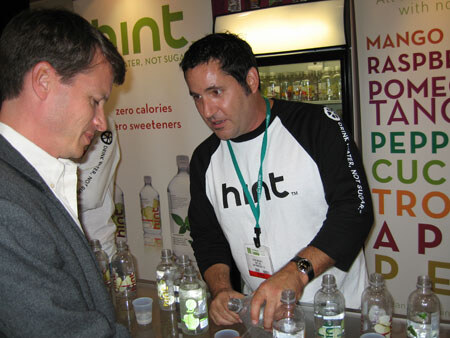 A man in a hint baseball shirt and lanyard pours from a bottle at a counter lined with hint bottles, while another man looks on. A flavor list is on the banner behind.