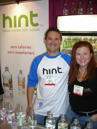 A man in a Hint baseball shirt and a woman with long red hair in a black Hint shirt smile at a booth. A Hint banner reads drink water, not sugar, and bottles sit on the counter.