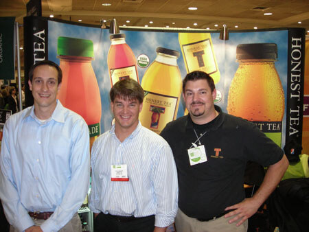 Three men stand in front of an Honest Tea backdrop showing large illustrated tea bottles. Two wear name badges, and the man on the right wears a dark polo shirt.