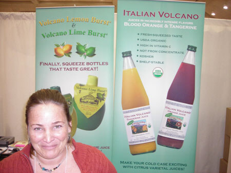 A smiling woman in a coral sweater stands in front of banners for Italian Volcano juices and Volcano Lemon Burst and Volcano Lime Burst squeeze bottles.