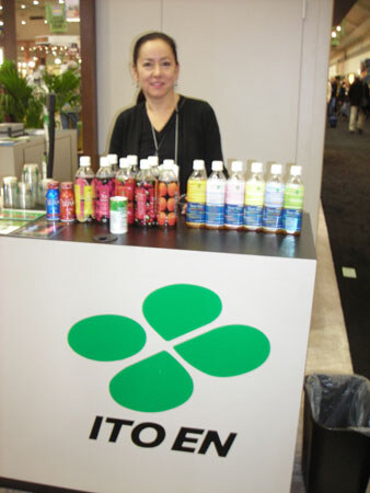 A smiling woman with dark hair stands behind a white Ito En counter with a green four-leaf logo. Rows of bottled and canned tea drinks sit on the counter in front of her.