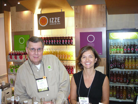 An older man in glasses and a woman in a black top stand smiling in front of an IZZE Sparkling Juice display with shelves and a cooler full of bottles.