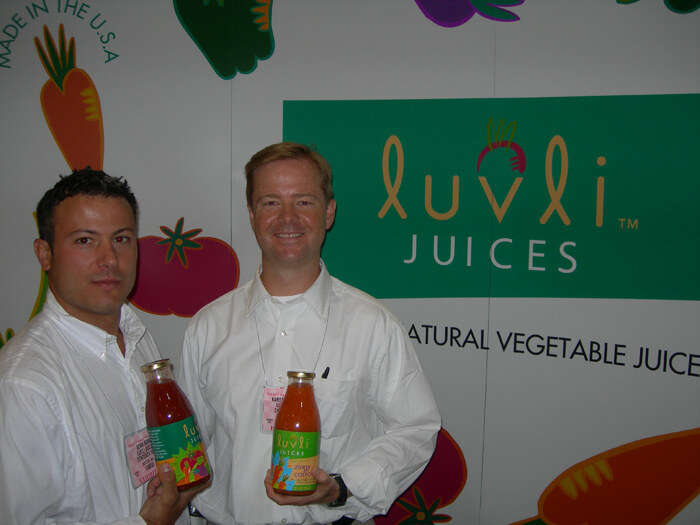 Two men in white shirts hold bottles of Luvli Juices vegetable juice in front of a green Luvli Juices sign and a backdrop of illustrated vegetables.