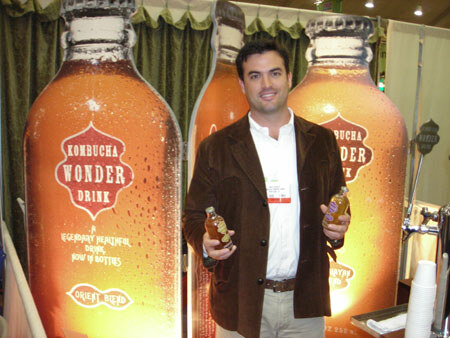A man in a brown jacket holds two small bottles in front of large Kombucha Wonder Drink bottle graphics, with a tap and stack of cups at right.