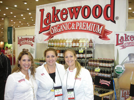 Three smiling women in white Lakewood jackets and badges stand in front of a Lakewood Organic & Premium banner and shelves of bottled juices.