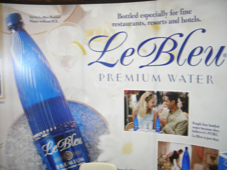 A Le Bleu Premium Water poster showing a blue bottle on ice, the text 'Bottled especially for fine restaurants, resorts and hotels,' and a photo of two people at a table.