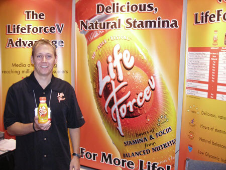 A smiling man in a black shirt holds a small bottle of LifeForceV in front of orange banners reading Delicious, Natural Stamina and The LifeForceV Advantage.