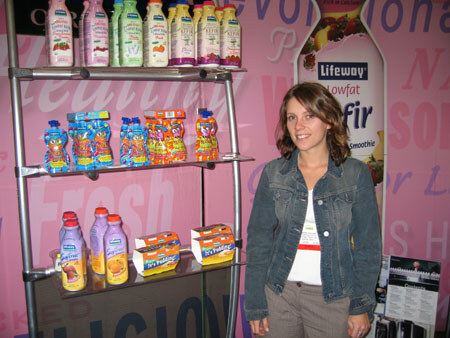 A woman in a denim jacket stands beside a metal shelf of Lifeway kefir bottles, pouches and pudding cups, with a pink backdrop and a Lifeway Lowfat Kefir banner behind her.