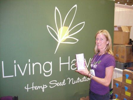 A smiling woman with blonde hair and a purple shirt holds a small carton in front of a green Living Harvest sign that reads Hemp Seed Nutrition. Boxes are stacked behind her.