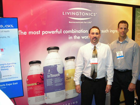 Two men in collared shirts and name badges stand in front of a Livingtonics display showing ResveraCarn, InTouch and ZenBrew bottles, with a screen to the left.
