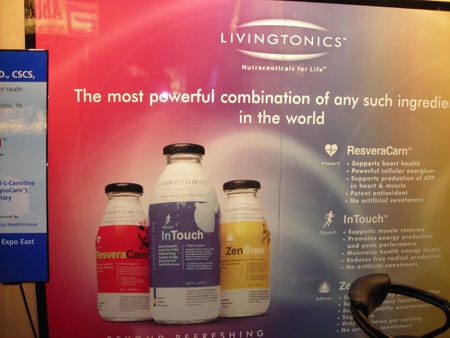 A Livingtonics sign shows bottles of ResveraCarn, InTouch and ZenBrew with text about each drink, beside a partly visible screen at the booth.