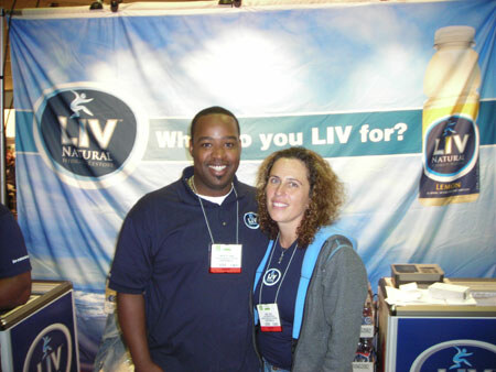 A man in a navy polo and a woman in a gray jacket, both wearing badges, stand together in front of a LIV Natural banner reading Who do you LIV for? with a lemon bottle pictured.