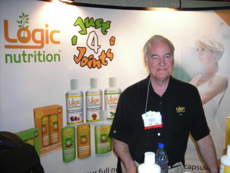 A man in a black Logic Nutrition shirt stands in front of a backdrop showing Logic Nutrition and Just 4 Joints bottles and boxes, with a bottle of water in front of him.
