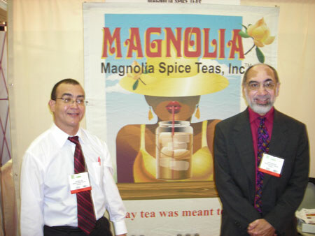Two men with name badges stand in front of a Magnolia Spice Teas, Inc. sign showing an illustration of a woman with a glass jar of iced tea and a straw.