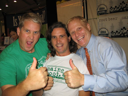 Three smiling men pose closely together, two giving thumbs up. One wears a Maine Root shirt, and a root beer banner hangs behind them.