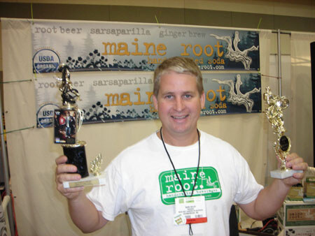 A smiling man in a white Maine Root t-shirt holds up two trophies in front of Maine Root banners listing root beer, sarsaparilla and ginger brew.