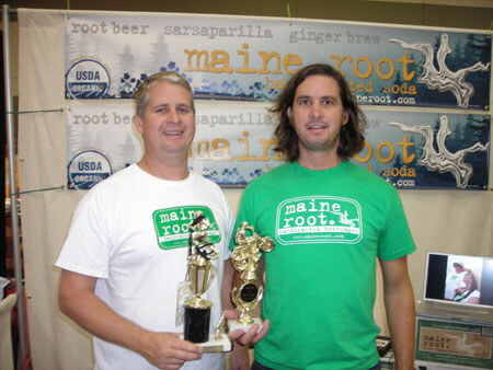 Two men in Maine Root t-shirts, one white and one green, stand together holding trophies in front of Maine Root banners at a trade show booth.