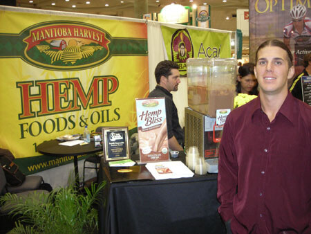 A man in a maroon shirt stands at a booth with a Manitoba Harvest Hemp Foods & Oils banner and a Hemp Bliss carton on a table; another man stands behind, and a beverage dispenser sits nearby.