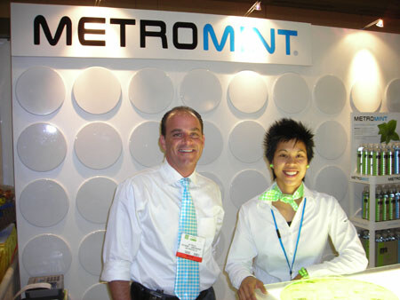 A smiling man in a white shirt and blue patterned tie stands beside a woman in a white jacket and green scarf in front of a large Metromint sign, with bottles on a shelf at right.