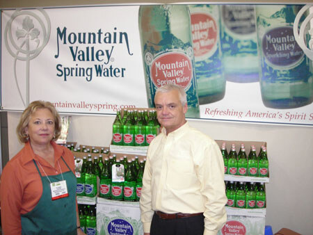 A woman in an apron and a man in a yellow shirt stand before a Mountain Valley Spring Water banner and cases of green glass bottles.