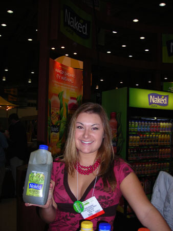 A smiling woman with a conference badge holds a large Naked juice bottle in front of a Naked-branded refrigerated display and signs at a trade show booth.