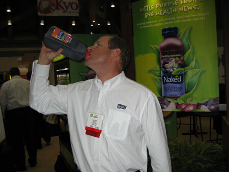 A man in a white Naked-branded shirt with a name badge tips back a dark bottle of juice to drink from it, in front of a Naked Purple Machine banner.