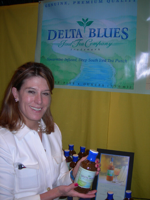 A smiling woman in a white jacket holds a bottle of Delta Blues spearmint-infused iced tea punch in front of a Delta Blues Iced Tea Company banner and more bottles on a table.