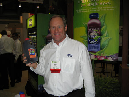 A smiling man in a white Naked-branded shirt and name badge holds up a dark bottle of Naked juice beside a large Naked Purple Machine banner.