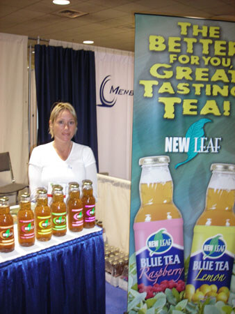 A blonde woman in a white top stands behind a blue-draped table with amber bottles of tea. A tall New Leaf banner shows Blue Tea Raspberry and Lemon bottles beside the words The Better For You Tea.
