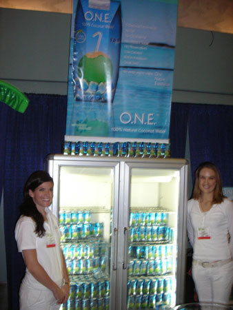 Two women in white stand on either side of a glass-door cooler stocked with O.N.E. coconut water cans. A banner above reads O.N.E. 100% Natural Coconut Water.