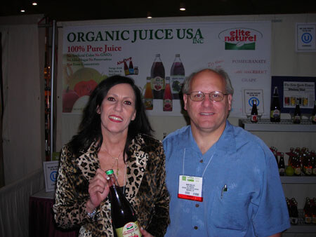 A woman in a leopard-print jacket holding a bottle of juice stands beside a man in glasses and a blue shirt in front of an Organic Juice USA Inc. and Elite Naturel sign.