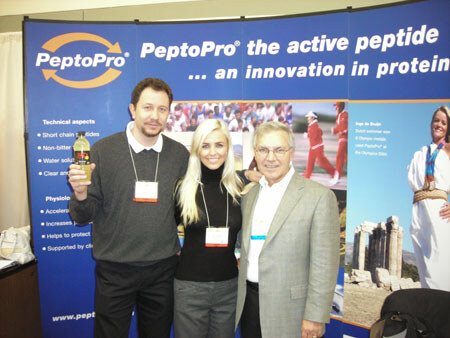 Two men and a blonde woman wearing badges pose in front of a blue PeptoPro banner reading the active peptide. The man on the left holds a bottle of amber drink.
