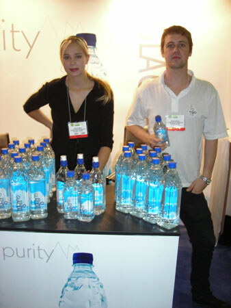 A blonde woman in black and a man in a white polo stand behind a table of Purity water bottles with blue caps, with Purity signage visible behind and in front of them.