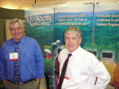 Two men wearing name badges stand in front of a Redeem Organic banner about 100% organic juices, with a green hillside and farm scene.
