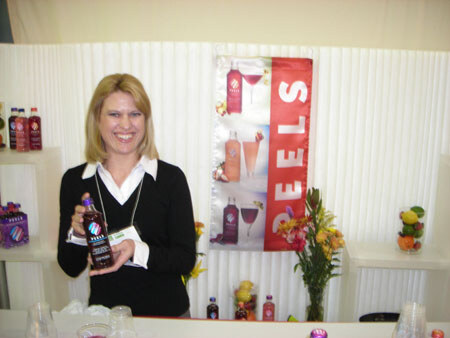 A smiling blonde woman in a black top holds a dark bottle in front of a red Reels banner showing bottles and cocktail glasses, with flowers and fruit nearby.
