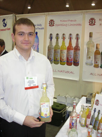 A young man in a white shirt holds a bottle of Rieme lemonade beside a table of bottles, with Rieme French Sparkling Limonades and sweetriot banners behind him.
