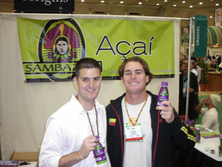 Two men smile in front of a green Sambazon Açaí banner, each holding a small bottle of drink. One wears a white shirt and the other a dark jacket with a name badge.