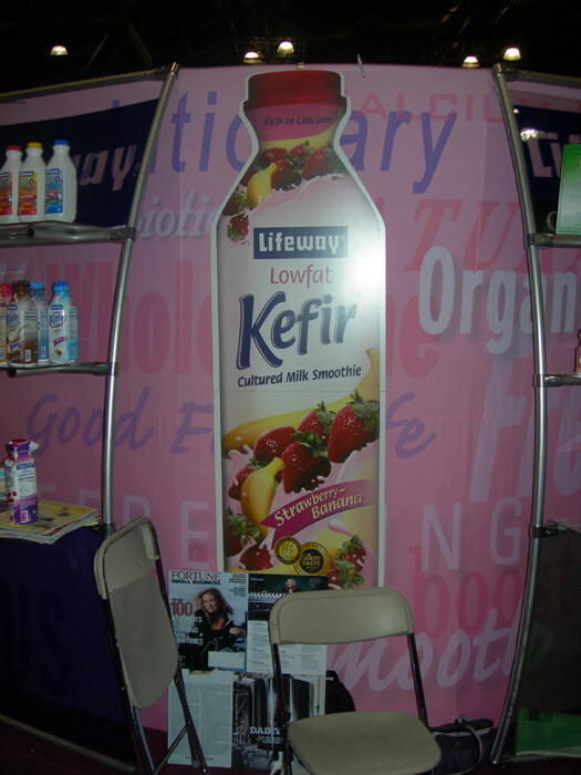 A pink Lifeway booth with a tall graphic of a Lowfat Strawberry-Banana Kefir bottle, shelves of kefir bottles on the left, and two folding chairs in front.