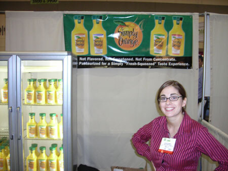 A smiling woman with glasses and a magenta striped shirt stands beside a refrigerated case of Simply Orange juice bottles, under a Simply Orange banner at a trade show booth.