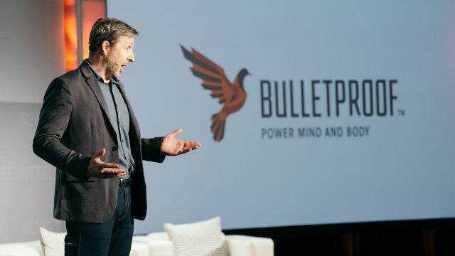 A man in a gray blazer with a headset microphone gestures on stage in front of a large screen showing the Bulletproof logo and the words Power Mind and Body.
