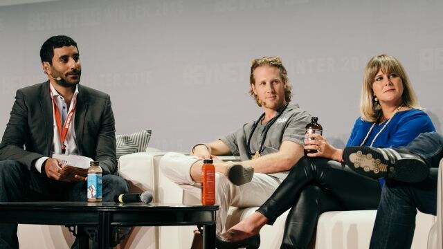 A man in a gray suit holds papers beside a table with drinks and a microphone. Next to him sit a man in a gray shirt and a woman in blue holding a Health-Ade Kombucha bottle.